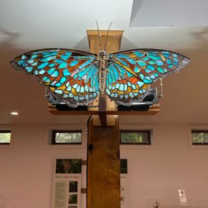 A metal sculpture of a butterfly with blue stained glass wings, illuminated and hanging from a wooden pillar in an art gallery