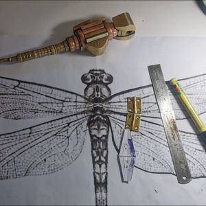 Paper designs of a dragonfly, with metal hinges and a steel ruler sitting on the paper. The sculpture of a large metal dragonfly sits on the paper