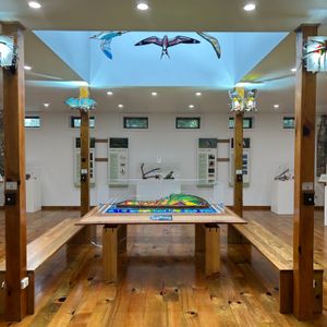 An indoor art gallery with a timber strip floor and white walls. In the centre of the room is a wooden table with a stained glass sculpture in the centre. On wooden pillars are sculptures of flying animals made of stained glass and metal