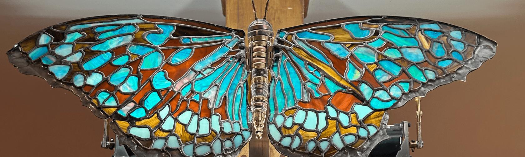 A metal sculpture of a butterfly with blue stained glass wings, illuminated and hanging from a wooden pillar in an art gallery