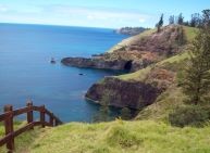 Looking out over rolling green hills and rocky cliffs against the sea. A wooden fence is in the foreground.